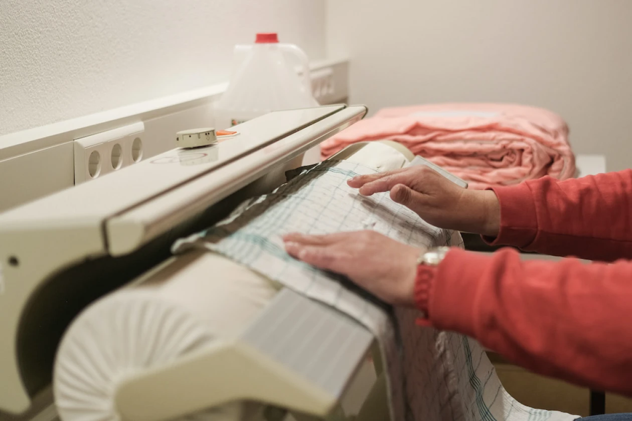 Blick auf eine B&uuml;gelmaschine: Zwei H&auml;nde einer Person im roten Pullover bedienen die Maschine und b&uuml;geln ein Geschirrtuch.