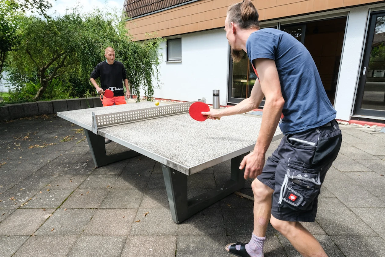 Zwei M&auml;nner spielen Tischtennis im Freien. Im Hintergrund ist ein Geb&auml;ude mit einer offenen Eingangst&uuml;r zu sehen.