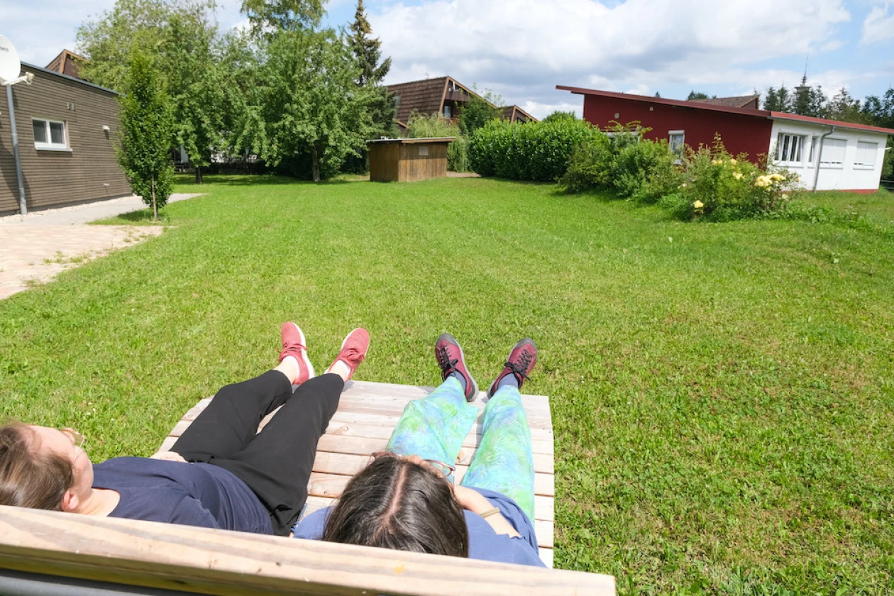 Zwei Personen liegen auf einer breiten Holzliege und genie&szlig;en den Blick auf die gr&uuml;ne Wiese und die umliegenden Geb&auml;ude.