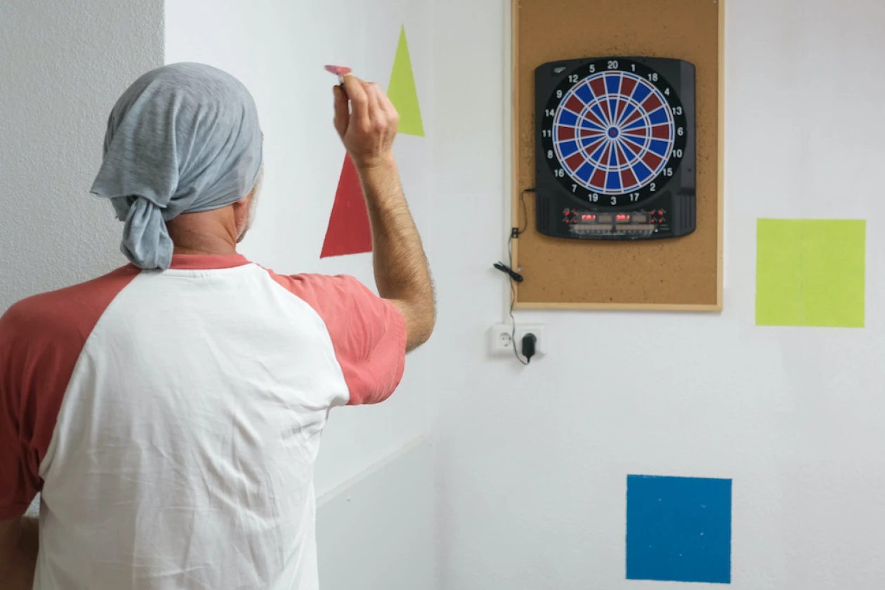 Zu sehen ist eine Person, die Dart spielt. An der Wand sind zwei Rechtecke und zwei Dreiecke in unterschiedlichen Farben abgebildet.