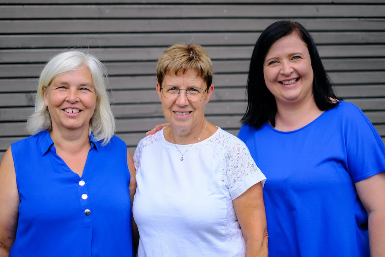 Drei Frauen l&auml;cheln direkt in die Kamera. Die Frau in der Mitte tr&auml;gt ein wei&szlig;es T-Shirt, w&auml;hrend die beiden anderen ein blaues Oberteil tragen.