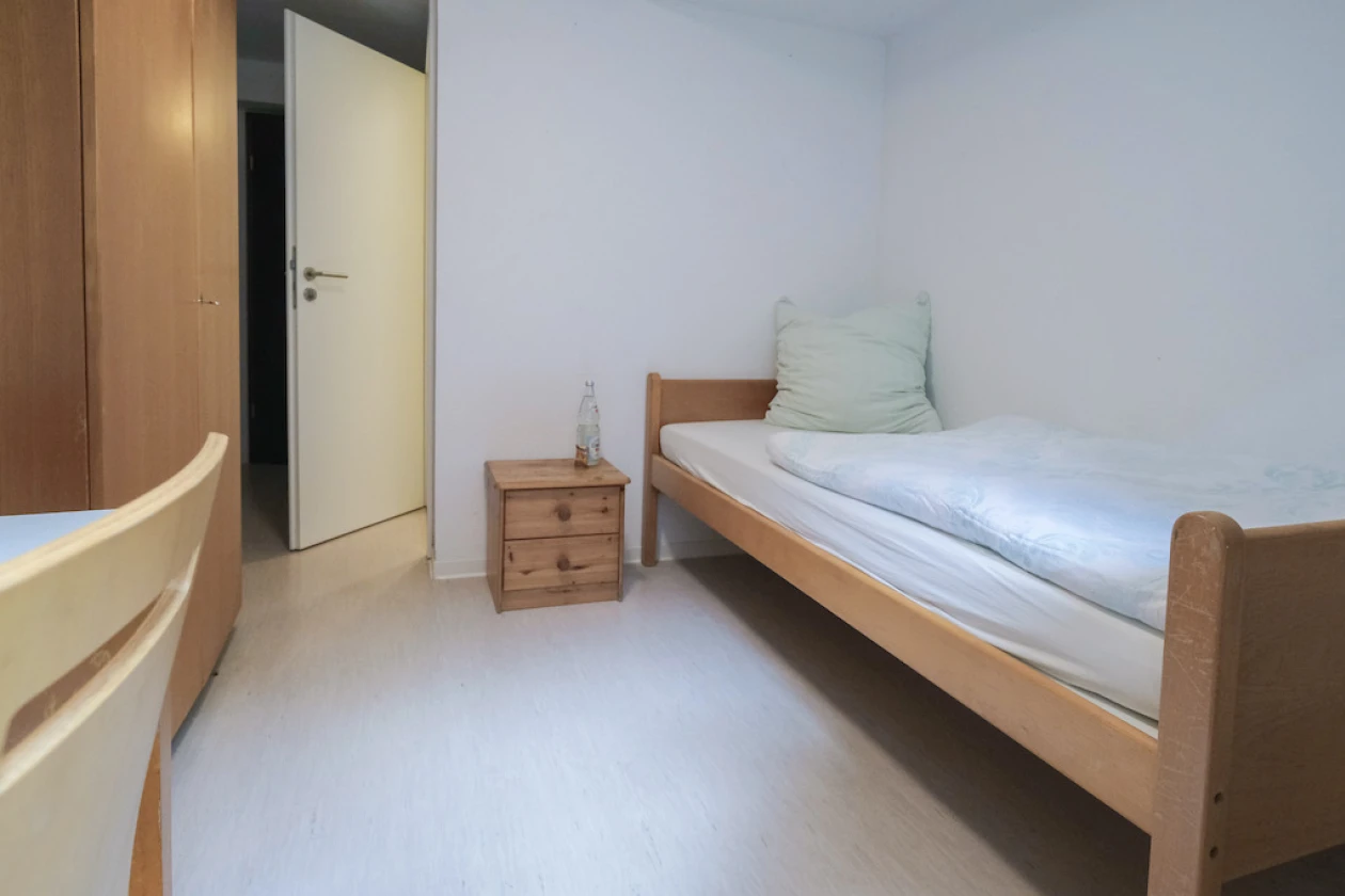 Ein Raum, der mit einem Holzbett, Nachttisch, einem Schrank, einem Tisch und einem Stuhl ausgestattet ist. Im Hintergrund ist eine ge&ouml;ffnete wei&szlig;e T&uuml;re zu sehen.