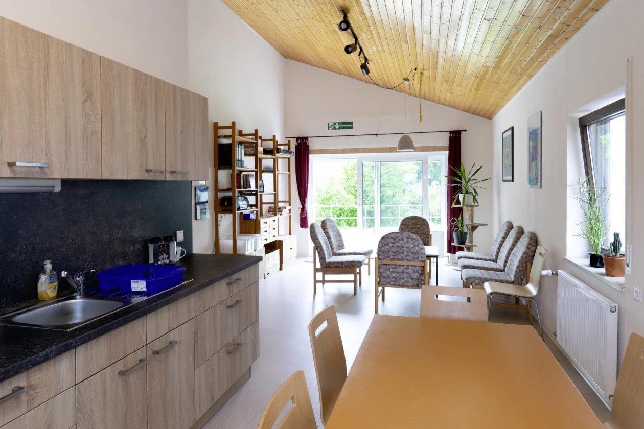 Zu sehen ist der Blick in eine Wohnk&uuml;che mit K&uuml;chenzeile, Esstisch und St&uuml;hlen und eine Sitzgruppe mit Regal im Hintergrund. chaftsraum mit K&uuml;che, Holztisch, St&uuml;hlen und einem Regal.