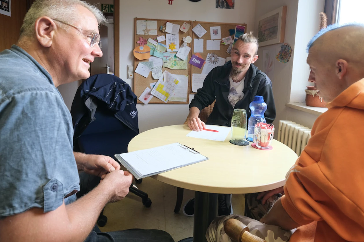 Drei M&auml;nner sitzen an einem runden Tisch. Der Mann links im Vordergrund spricht, die anderen beiden h&ouml;ren zu. Auf dem Tisch liegen Dokumente, zwei Gl&auml;ser und eine Wasserflasche. Im Hintergrund ist eine Pinnwand zu sehen.