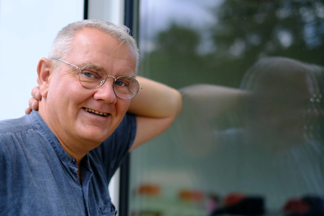 Ein Mann mit Brille und grauem Haar lehnt sich an ein Fenster und l&auml;chelt in die Kamera.