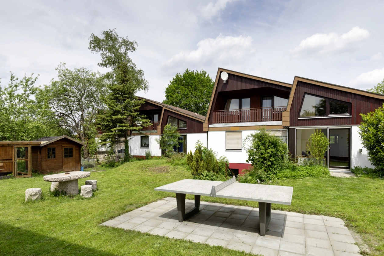 Vor einer Gruppe kleiner Wohnh&auml;user und einem Gartenh&auml;uschen ist eine Rasenfl&auml;che mit einer Tischtennisplatte zu sehen.