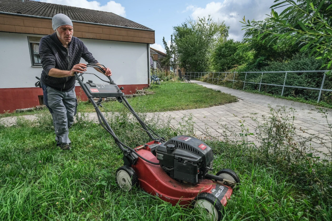 Ein Mann in Arbeitskleidung mit M&uuml;tze m&auml;ht mit einem roten Rasenm&auml;her das hohe Gras vor einem wei&szlig;en Geb&auml;ude.