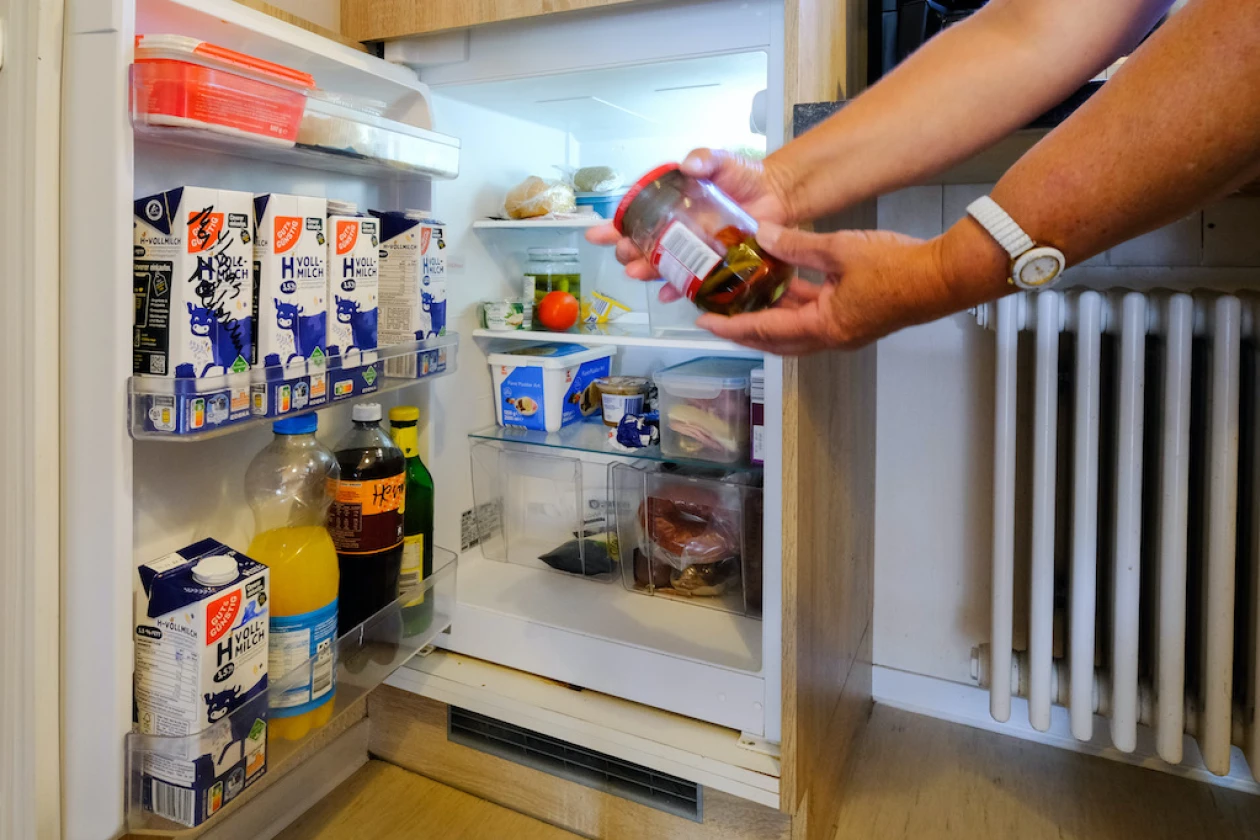 Ein K&uuml;hlschrank ist ge&ouml;ffnet, in dem verschiedene Milchprodukte, Getr&auml;nke und Lebensmittel stehen. Eine Hand greift nach einem Glas mit rotem Deckel. Der Inhalt ist unscharf und nicht zu erkennen.
