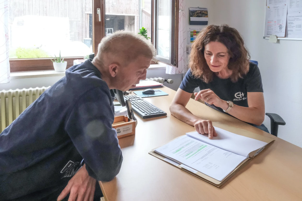 Eine Frau in einem dunkelblauen T-Shirt mit dem Logo der "Erlacher H&ouml;he" sitzt hinter dem Schreibtisch und zeigt mit dem Finger auf ein Dokument, w&auml;hrend der Mann vor dem Schreibtisch sitzt und interessiert zuh&ouml;rt. Im Hintergrund ist ein ge&ouml;ffnetes Fenster mit Blick ins Gr&uuml;ne zu sehen.