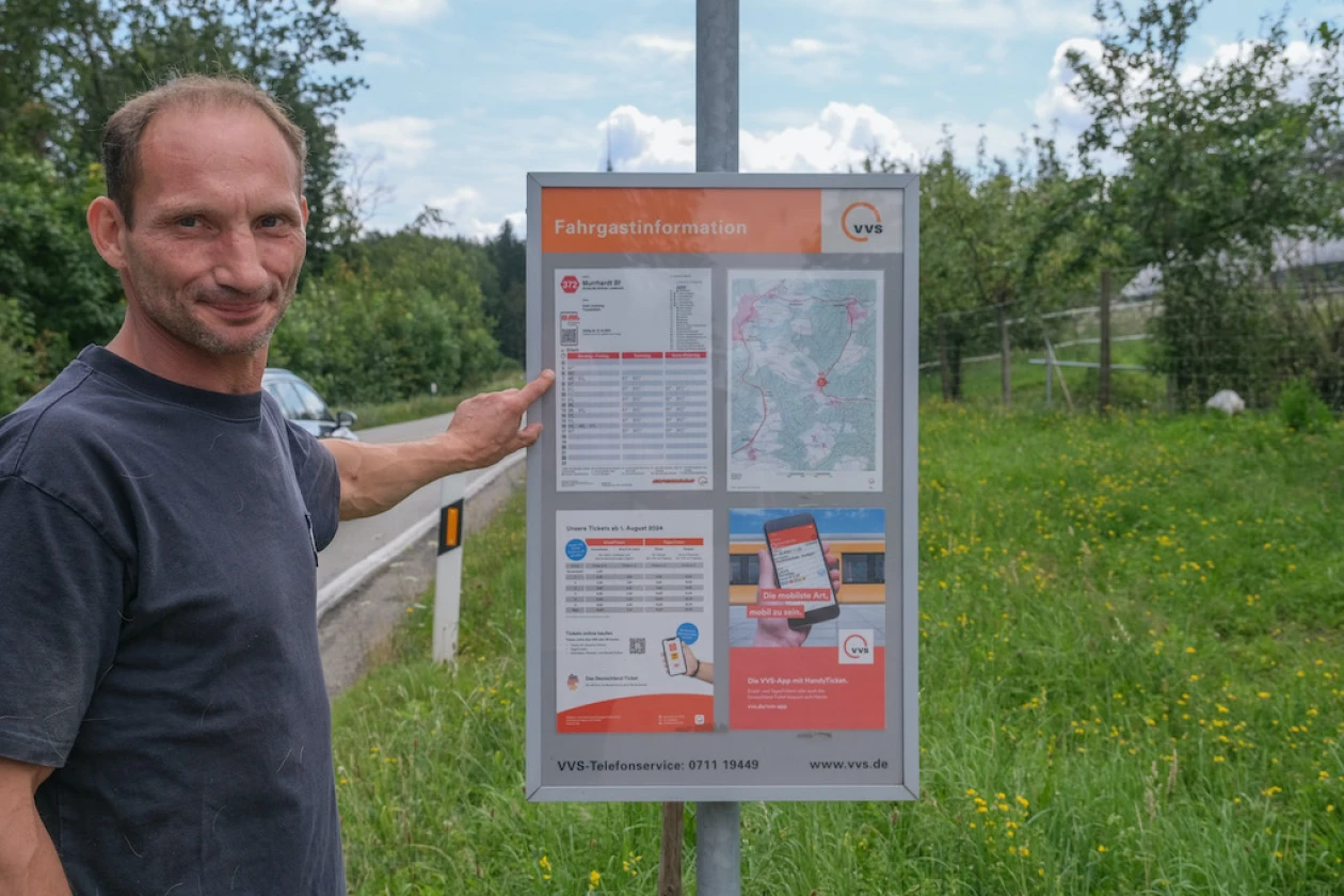 Ein Mann steht an einer Bushaltestelle und zeigt mit dem Finger auf eine Fahrplantafel. Im Hintergrund ist eine gr&uuml;ne Landschaft und ein vorbeifahrendes Auto zu sehen.