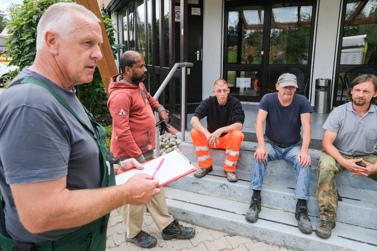 F&uuml;nf M&auml;nner sind zu sehen: Drei sitzen auf einer Treppe, w&auml;hrend zwei stehen. Einer der Stehenden h&auml;lt ein Dokument und einen Kugelschreiber in der Hand. Im Hintergrund ist ein Geb&auml;ude mit Glasfenstern erkennbar.