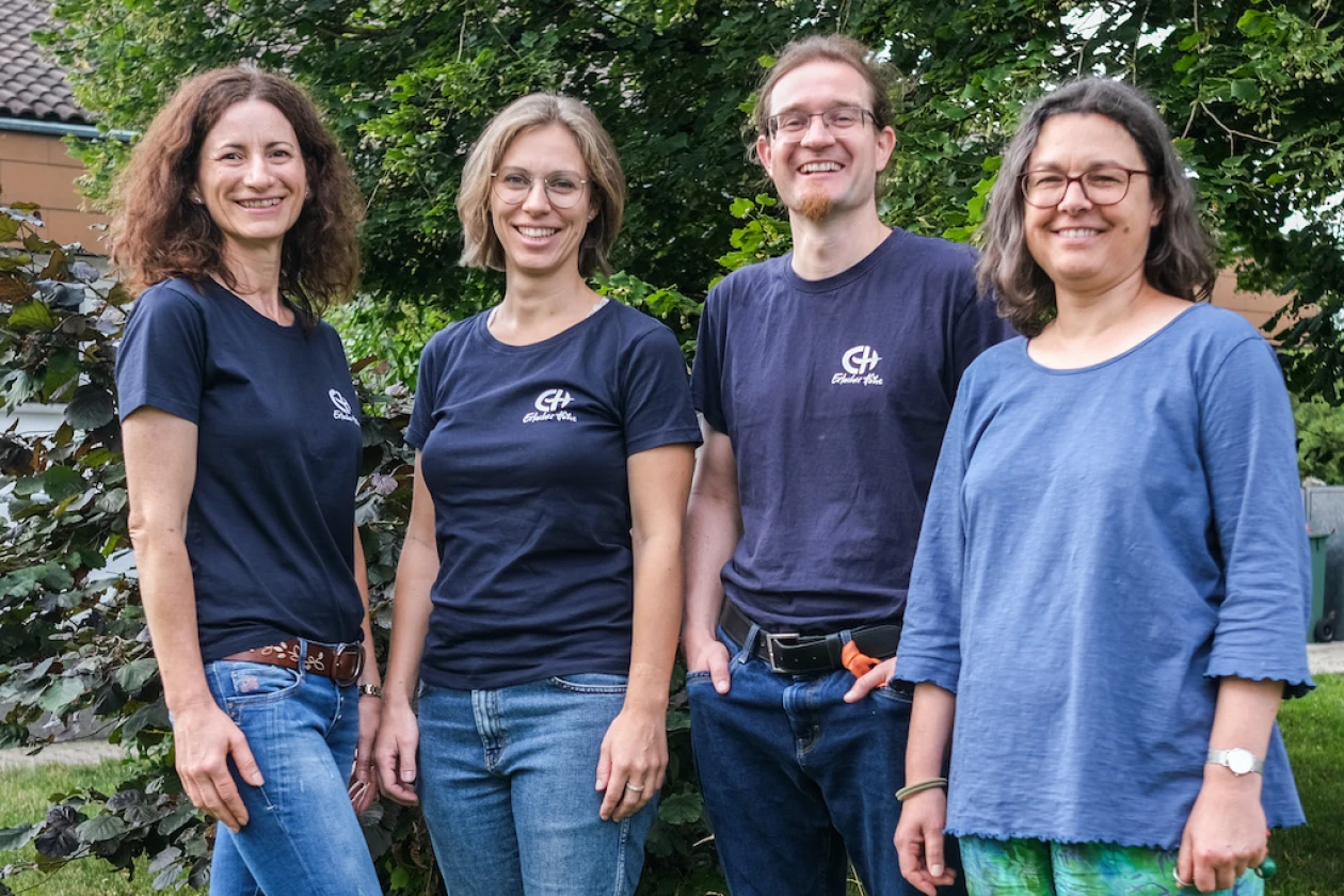 Drei Frauen und ein Mann l&auml;cheln in die Kamera, drei davon tragen ein dunkelblaues T-Shirt mit dem Logo der Erlacher H&ouml;he. Im Hintergrund sind B&auml;ume und Str&auml;ucher zu sehen.