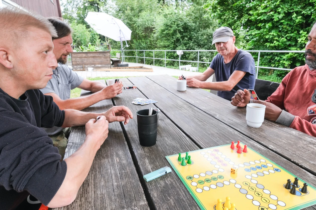 Vier Personen sitzen an einem Tisch, auf dem drei Tassen, Uno-Karten und das Brettspiel "Mensch &auml;rgere dich nicht" liegen.
