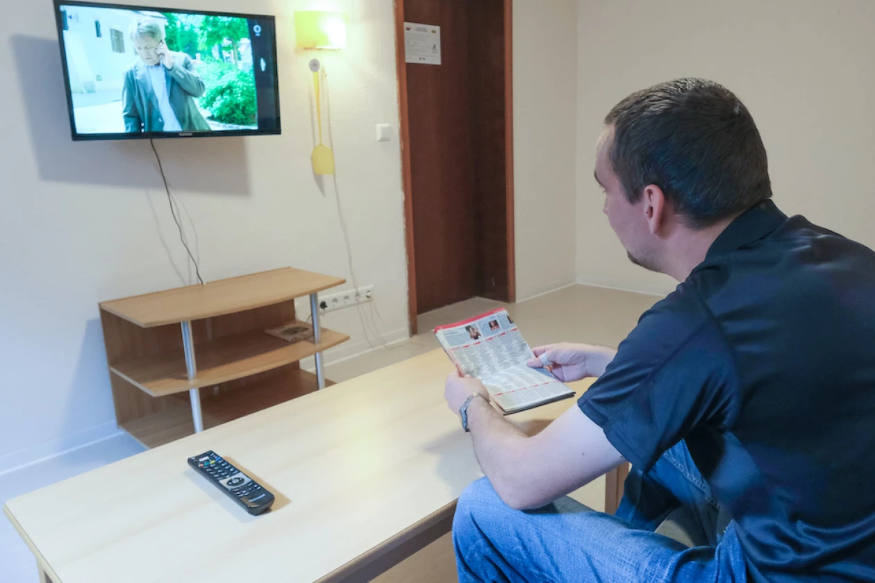 Ein Mann sitzt in einem Raum mit einem Holztisch, einem kleinen Regal und einem Fernseher an der Wand. Er h&auml;lt ein TV-Zeitschrift in der Hand.
