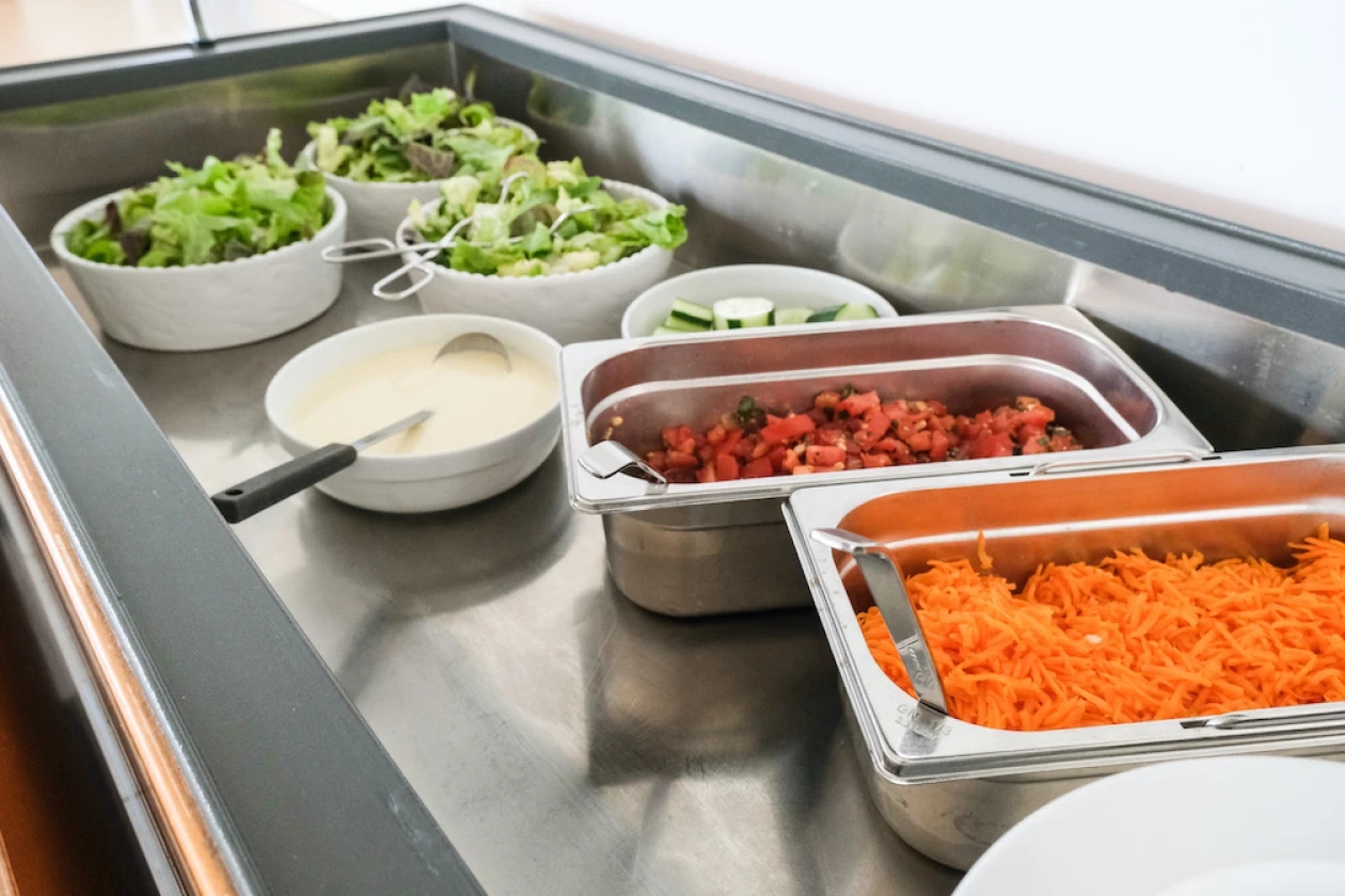Blick in ein Salatbuffet: Drei wei&szlig;e Schalen mit Salatbl&auml;ttern, eine Sch&uuml;ssel mit einer wei&szlig;en Sauce, eine Metallschale voller Tomaten und eine weitere Schale mit geriebenen M&ouml;hren sind zu sehen.