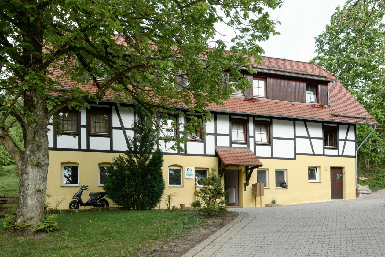 Fachwerkhaus mit gelbem Sockel am Untergeschoss und Fachwerk am Obergeschoss. Im rot eingedeckten Ziegeldach ist ist eine Gaube eingebaut. Das Haus ist umgeben von B&auml;umen und einer gepflasterten Einfahrt. Ein Motorroller ist seitlich vor dem Haus geparkt.