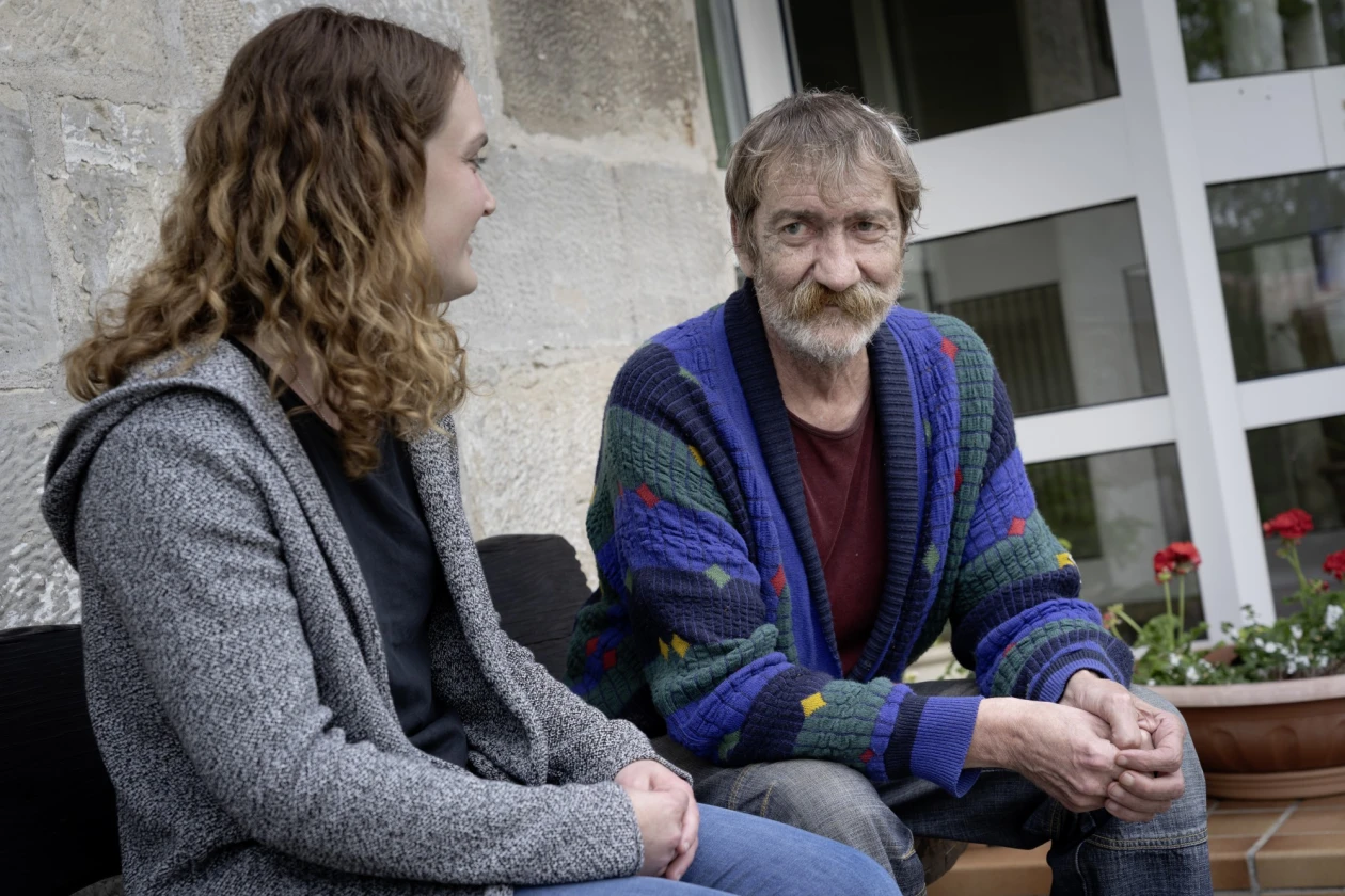 Ein Mann mit gr&uuml;n-blauer gestreifter Strickjacke sitzt neben einer Mitarbeiterin in grauer Jacke. Sie sitzen gemeinsam vor einem Fenster. Im Hintergrund ist ein Blumentopf zu sehen.