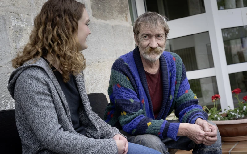 Ein Mann mit gr&uuml;n-blauer gestreifter Strickjacke sitzt neben einer Mitarbeiterin in grauer Jacke. Sie sitzen gemeinsam vor einem Fenster. Im Hintergrund ist ein Blumentopf zu sehen.