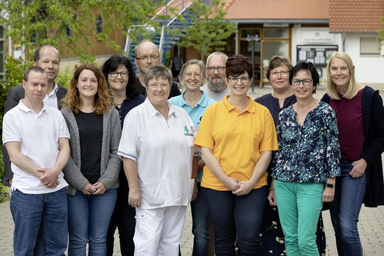 Zw&ouml;lf Personen unterschiedlichen Alters und Geschlechts l&auml;cheln direkt in die Kamera. Die Gruppe steht vor einem Geb&auml;ude mit B&auml;umen im Hintergrund.