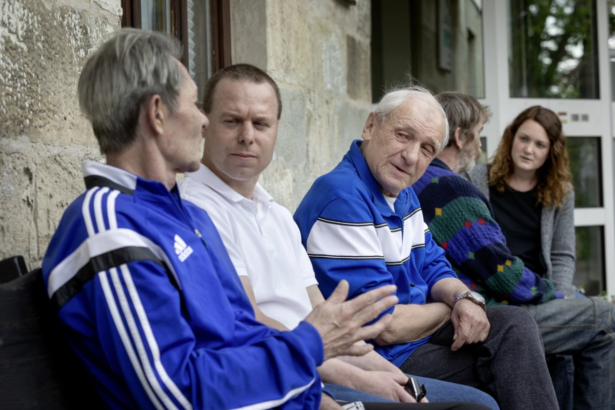 F&uuml;nf Personen sitzen drau&szlig;en vor einer Steinmauer und unterhalten sich.