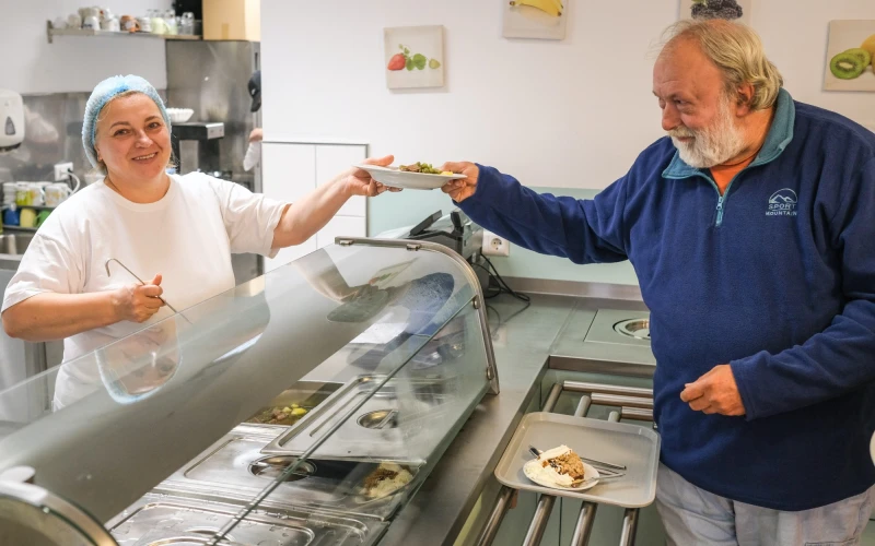 Eine Frau in wei&szlig;er Kleidung mit Haube l&auml;chelt und reicht einem Mann in blauer Jacke einen Teller Essen &uuml;ber eine Theke in einer Kantine.