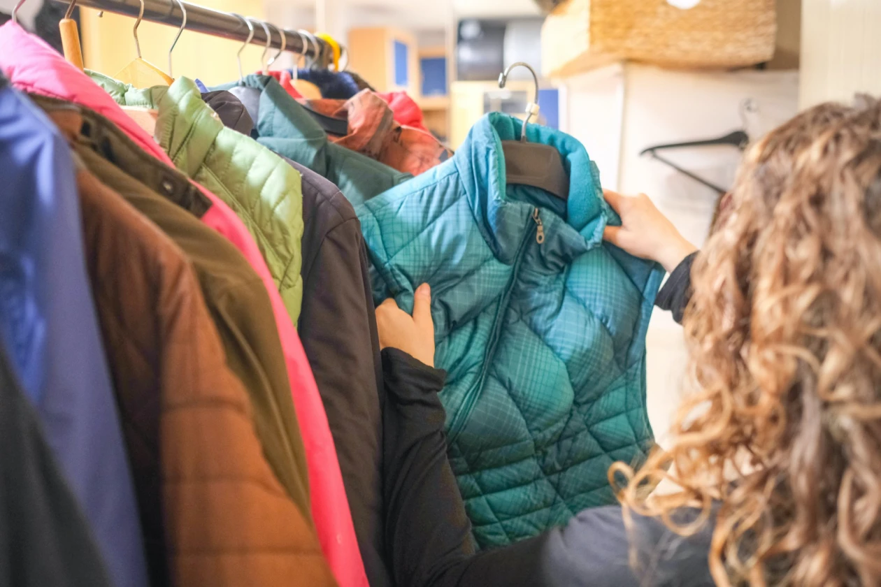 Winterjacken in verschiedenen Farben h&auml;ngen an einer Kleiderstange. Eine Person mit lockigen Haaren steht seitlich und betrachtet eine gr&uuml;ne Weste, die sie in den H&auml;nden h&auml;lt.
