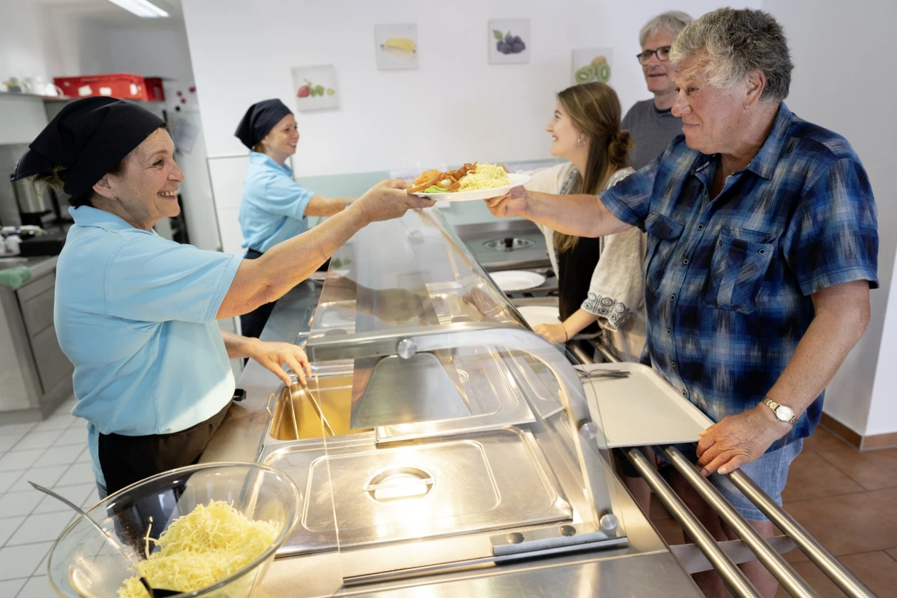 Zwei Frauen in hellblauen T-Shirts geben Essen an G&auml;ste an einer Ausgabetheke aus. Eine Frau reicht einem Mann in kariertem Hemd eine Portion Essen, w&auml;hrend im Hintergrund zwei weitere Personen warten.