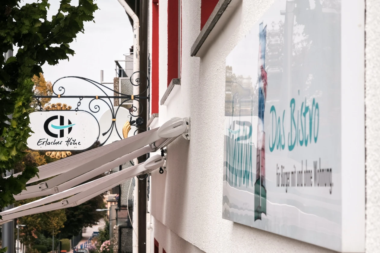 Metallschild an einem Geb&auml;ude mit wei&szlig;er Fassade und roten Fensterrahmen mit der Aufschrift "Erlacher H&ouml;he".