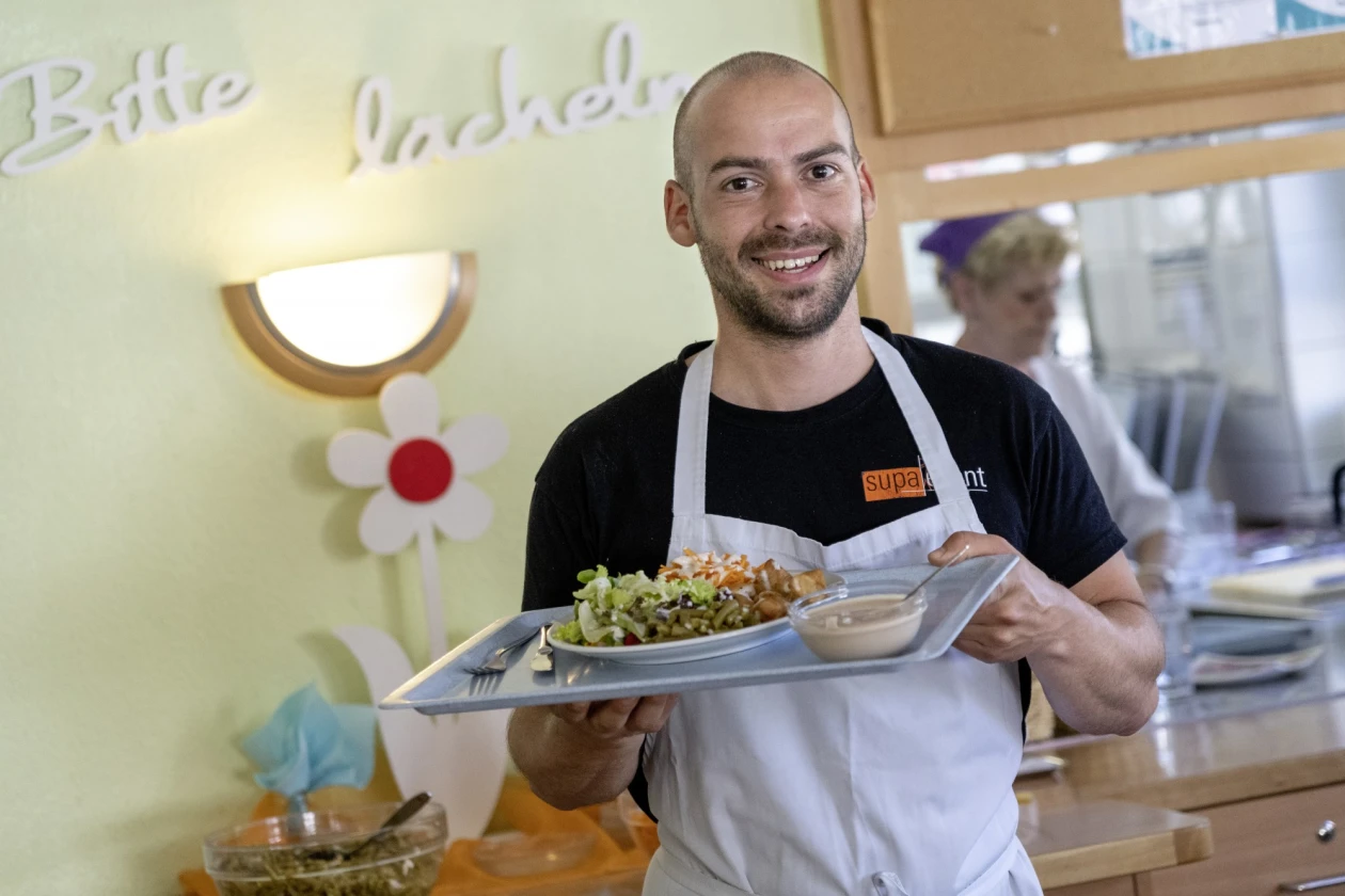 Ein Mann in wei&szlig;er Sch&uuml;rze l&auml;chelt in der K&uuml;che und pr&auml;sentiert ein Tablett mit einem Teller mit gr&uuml;nem Salat und einer Sch&uuml;ssel. Er steht vor einer pastellgelben Wand mit einer dekorativen wei&szlig;en Blume. Im Hintergrund ist eine weitere Person hinter der Theke zu sehen.
