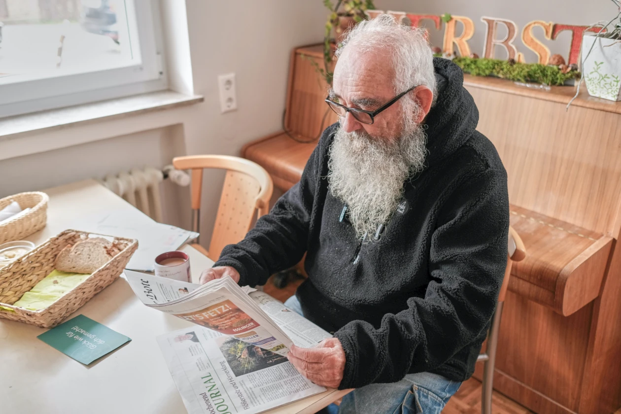 Ein Mann mit langem wei&szlig;em Bart und Brille sitzt an einem Tisch und liest Zeitung, w&auml;hrend Fr&uuml;hst&uuml;cksutensilien daneben stehen. Im Hintergrund ist ein Klavier zu sehen.