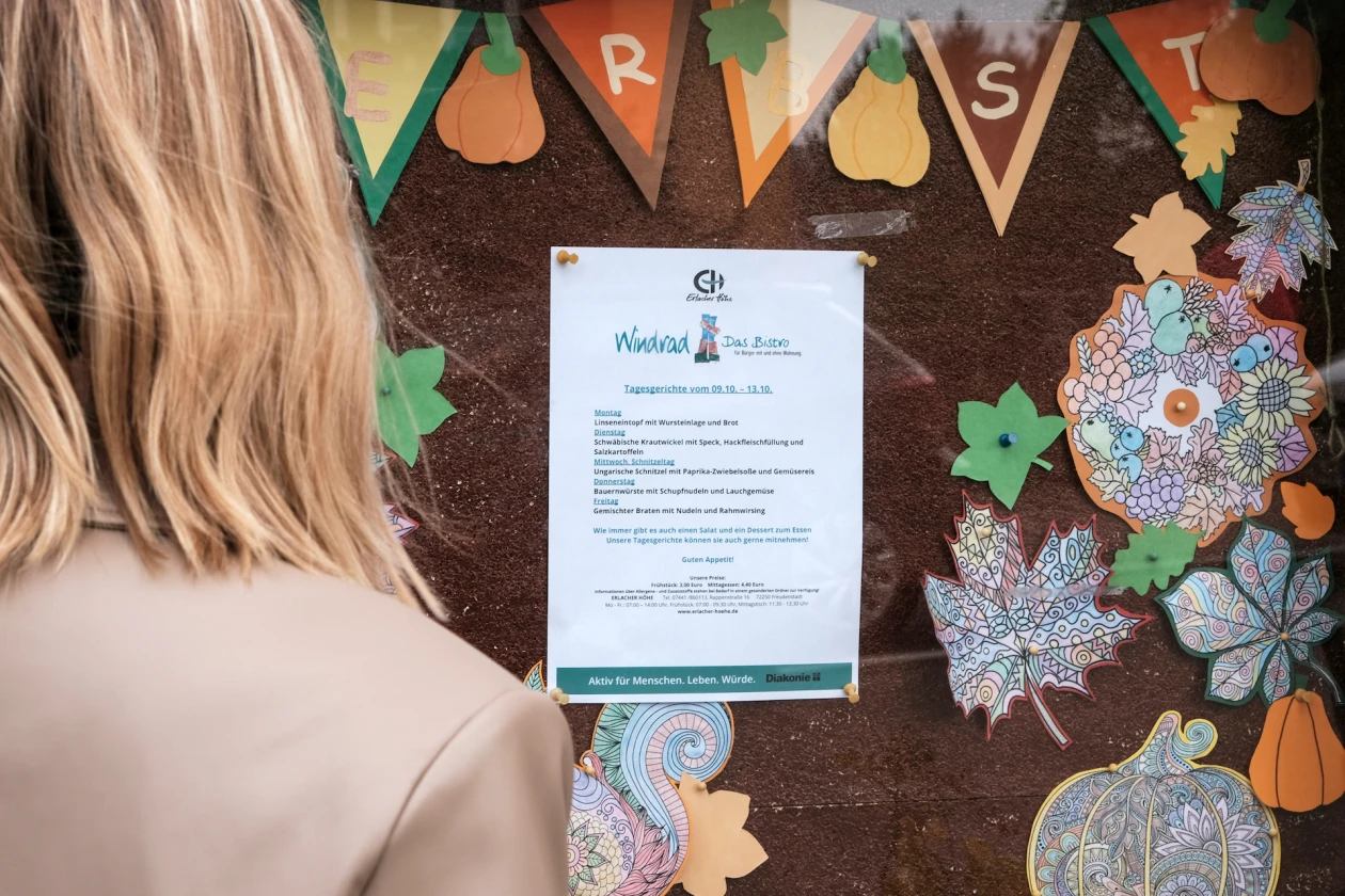 Eine Person steht vor einer Wand, an der ein Aushang befestigt ist. Auf dem Dokument steht "Windrad" das Bistro mit dem Logo der Erlacher H&ouml;he.