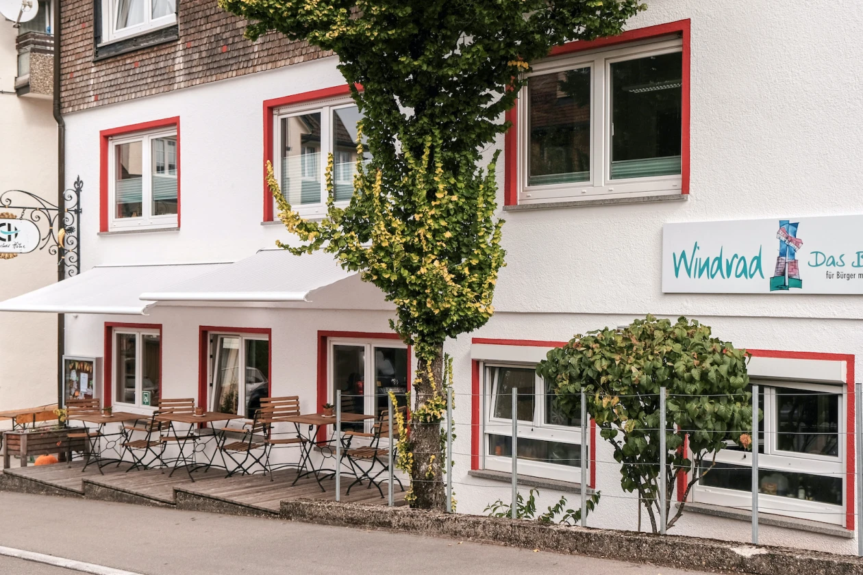 Wei&szlig;-rotes Geb&auml;ude mit Au&szlig;enbereich, bestehend aus Bistrotischen mit St&uuml;hlen, teilweise von einem Baum beschattet. An der Fassade h&auml;ngt ein Schild mit dem Schriftzug "Windrad" Das Bistro." Die Fenster haben rote Rahmen.