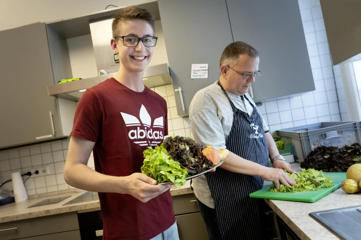 Zwei Personen in einer K&uuml;che, die frischen Salat zubereiten. Ein Mann in einem roten T-Shirt l&auml;chelt und h&auml;lt einen Teller mit Salat in der Hand, w&auml;hrend der andere Mann in einer gestreiften Sch&uuml;rze Salat auf einem gr&uuml;nen Schneidebrett schneidet.