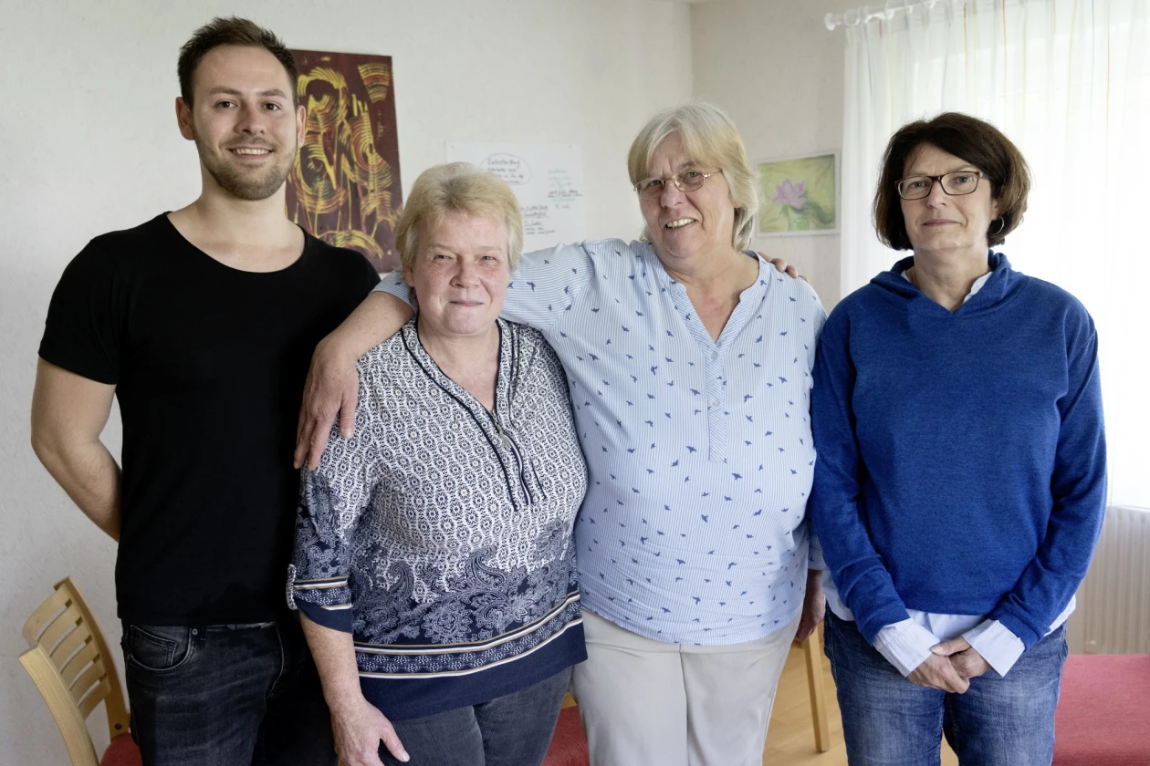 Vier Personen blicken direkt in die Kamera, sie stehen nebeneinander in einem Raum mit wei&szlig;en W&auml;nden, wei&szlig;en Vorh&auml;ngen am Fenster und Holzst&uuml;hlen. Im Hintergrund sind Bilder an der Wand aufgeh&auml;ngt.