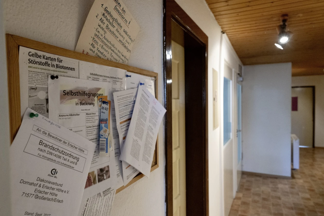 Pinnwand mit verschiedenen angehefteten Dokumenten in einem Flur mit Holzdecke und T&uuml;ren. An der Decke brennt ein Licht.