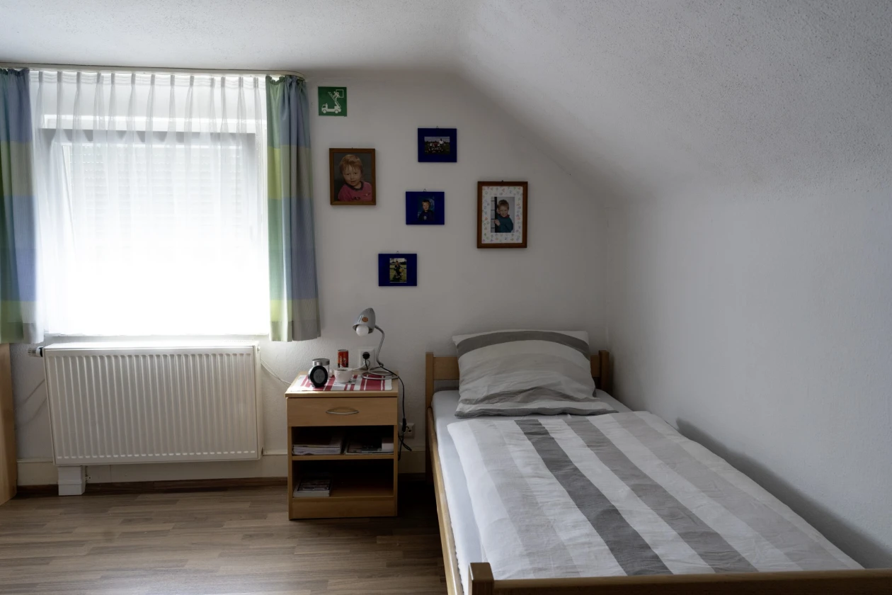 Ein gem&uuml;tliches Zimmer mit einem Bett samt gestreiftem Bettbezug, einem kleinen Nachttisch aus Holz und einem wei&szlig;en Heizk&ouml;rper. Am Fenster h&auml;ngen wei&szlig;e Vorh&auml;nge, und mehrere gerahmte Bilder schm&uuml;cken die Wand &uuml;ber dem Bett.