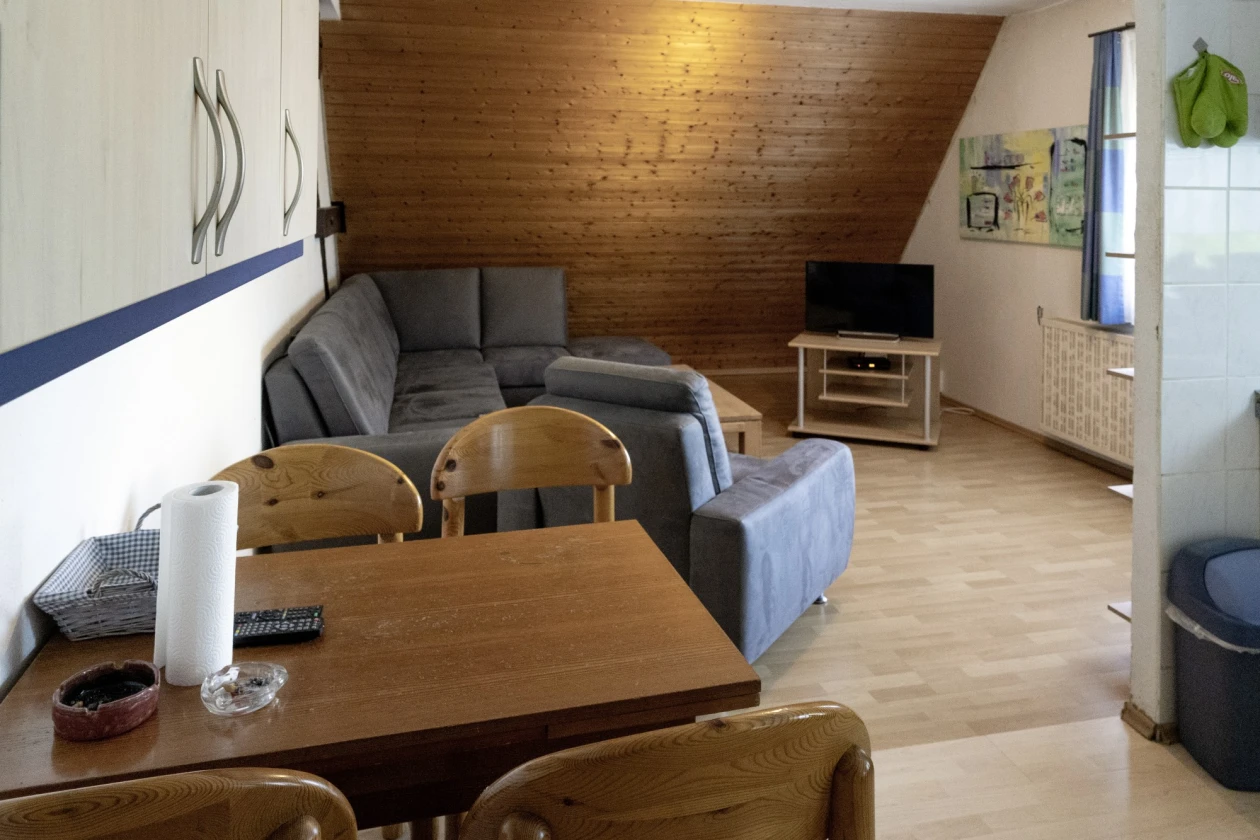 Ein gem&uuml;tlich eingerichtetes Appartement mit einem grauen Ecksofa, Fernseher und einem Holztisch vier St&uuml;hlen. Die Dachschr&auml;ge im Raum ist mit Holz verkleidet. Ein Fenster und ein Gem&auml;lde ist im Hintergrund zu sehen. Der Raum ist mit Parkettboden und wei&szlig;en W&auml;nden gestaltet.