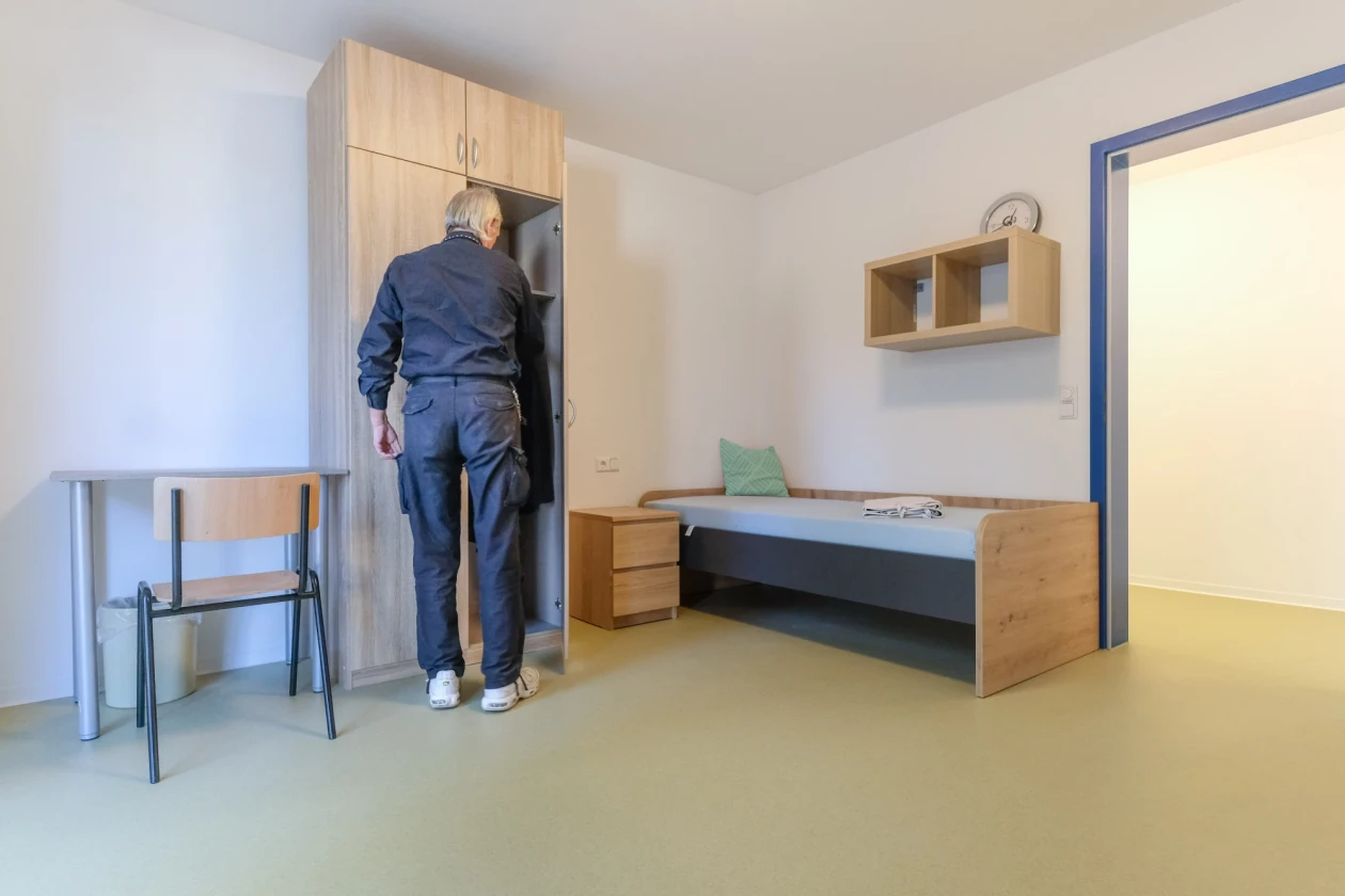 Mann in dunkler Kleidung steht vor offenem Schrank in schlicht eingerichtetem Zimmer mit Bett, Tisch, Stuhl und Wandregal.