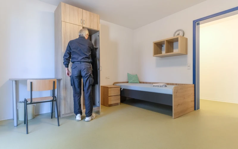 Mann in dunkler Kleidung steht vor offenem Schrank in schlicht eingerichtetem Zimmer mit Bett, Tisch, Stuhl und Wandregal.