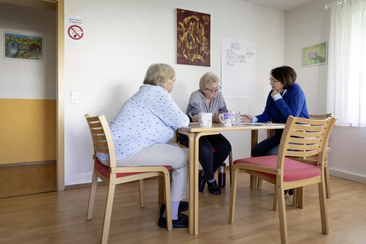 Drei Frauen sitzen an einem Holztisch in einem hellen Raum mit Bildern an den W&auml;nden und einem Fenstern mit wei&szlig;en Vorh&auml;ngen. Auf dem Tisch stehen Tassen. Sie unterhalten sich.