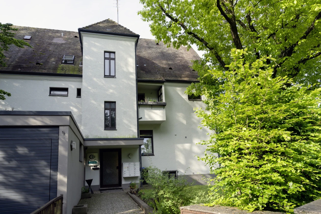 Ein gro&szlig;es, wei&szlig;es Geb&auml;ude mit Garage, umgeben von &uuml;ppigem, gr&uuml;nem Laubbaum. Der moderne und gepflegte Eingangsbereich verf&uuml;gt &uuml;ber eine Eingangst&uuml;r sowie einen gepflasterten Weg. Neben der Eingangst&uuml;r ist eine Briefkastenanlage und das Logo der Erlacher H&ouml;he angebracht.