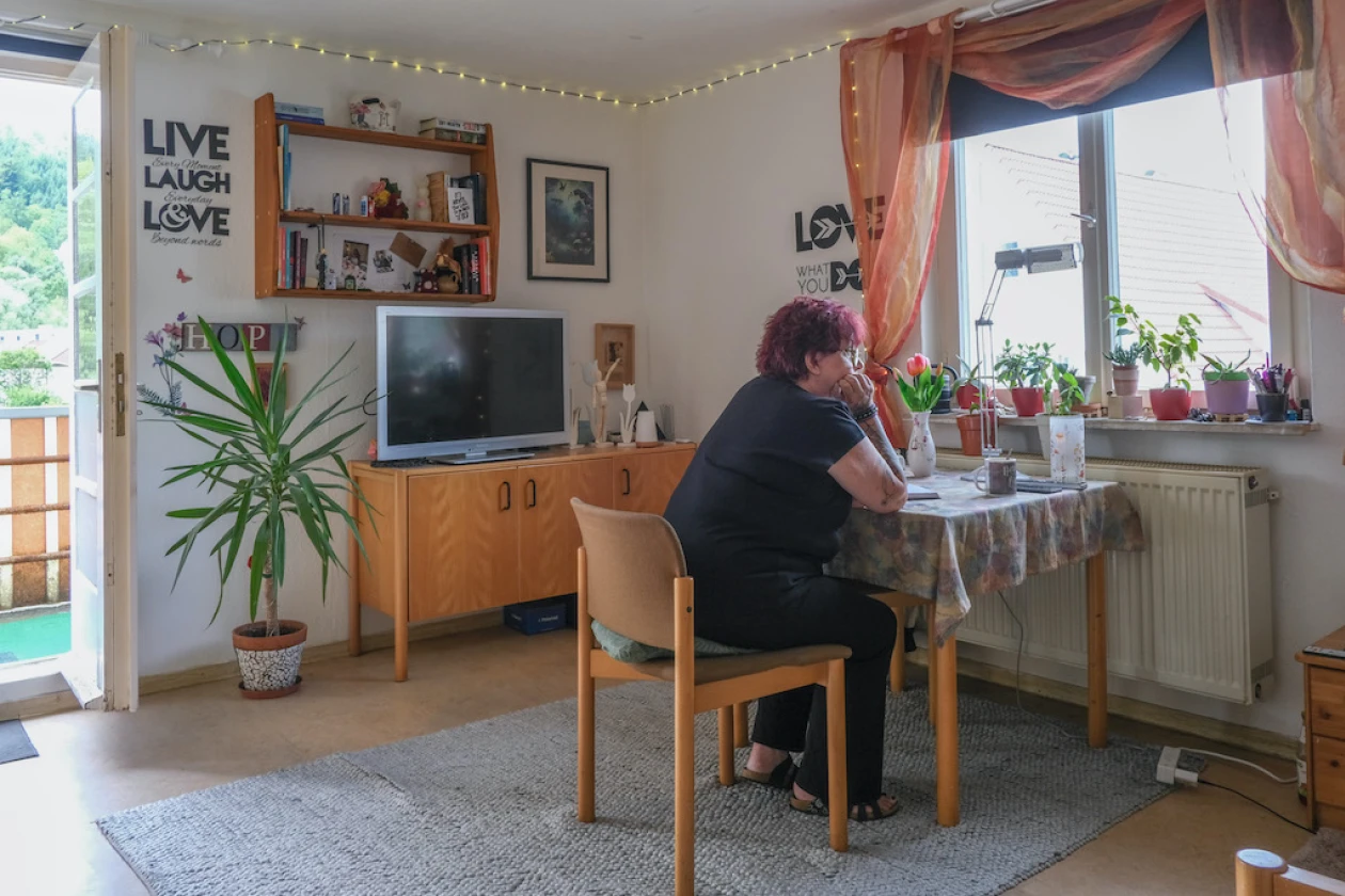 Eine Frau sitzt an einem Holztisch in einem hellen Wohnzimmer und blickt nachdenklich aus dem Fenster. Im Raum befinden sich ein Schrank mit einem Fernsehger&auml;t, ein B&uuml;cherregal und mehrere Zimmerpflanzen. An der Wand h&auml;ngen Lichterketten sowie dekorative Schriftz&uuml;ge wie "Live Laugh Love". Vor dem Fenster h&auml;ngen rote Vorh&auml;nge.