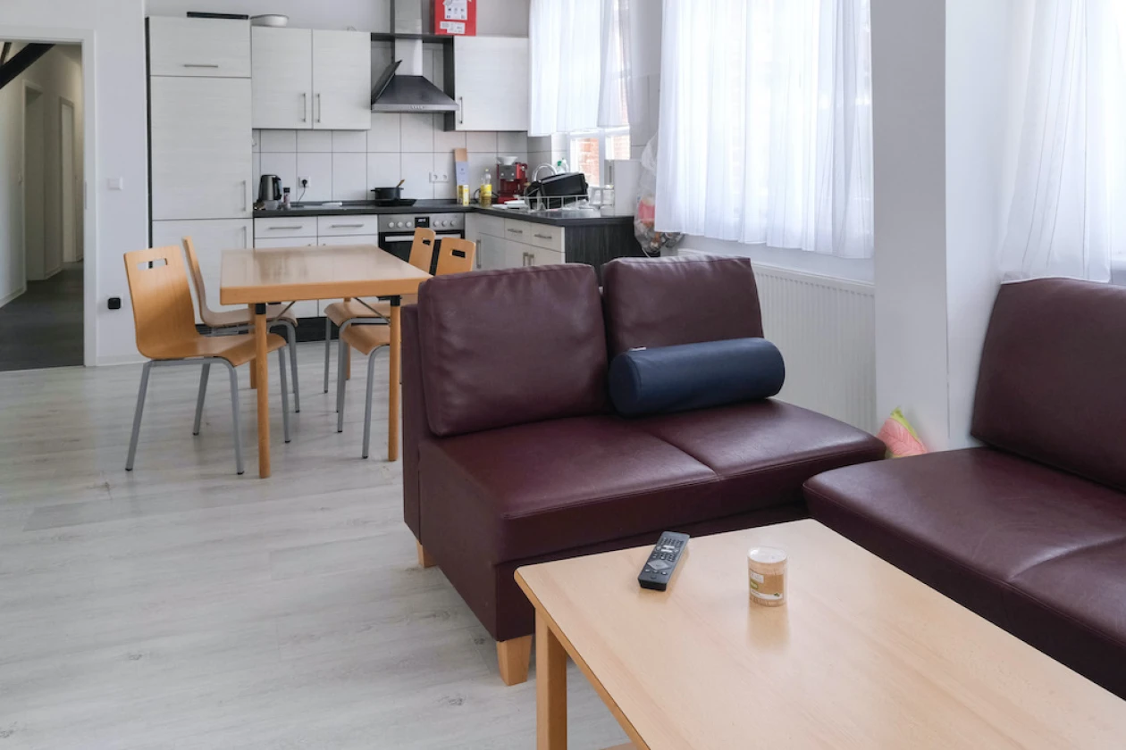 Ein Raum mit offener K&uuml;che, Essbereich mit Holztisch und Holzst&uuml;hlen, sowie Wohnbereich mit roten Ledersofas und Holztisch. Die wei&szlig;en W&auml;nde und Fenster mit wei&szlig;en Vorh&auml;ngen verleihen dem Raum eine helle, freundliche Atmosph&auml;re.