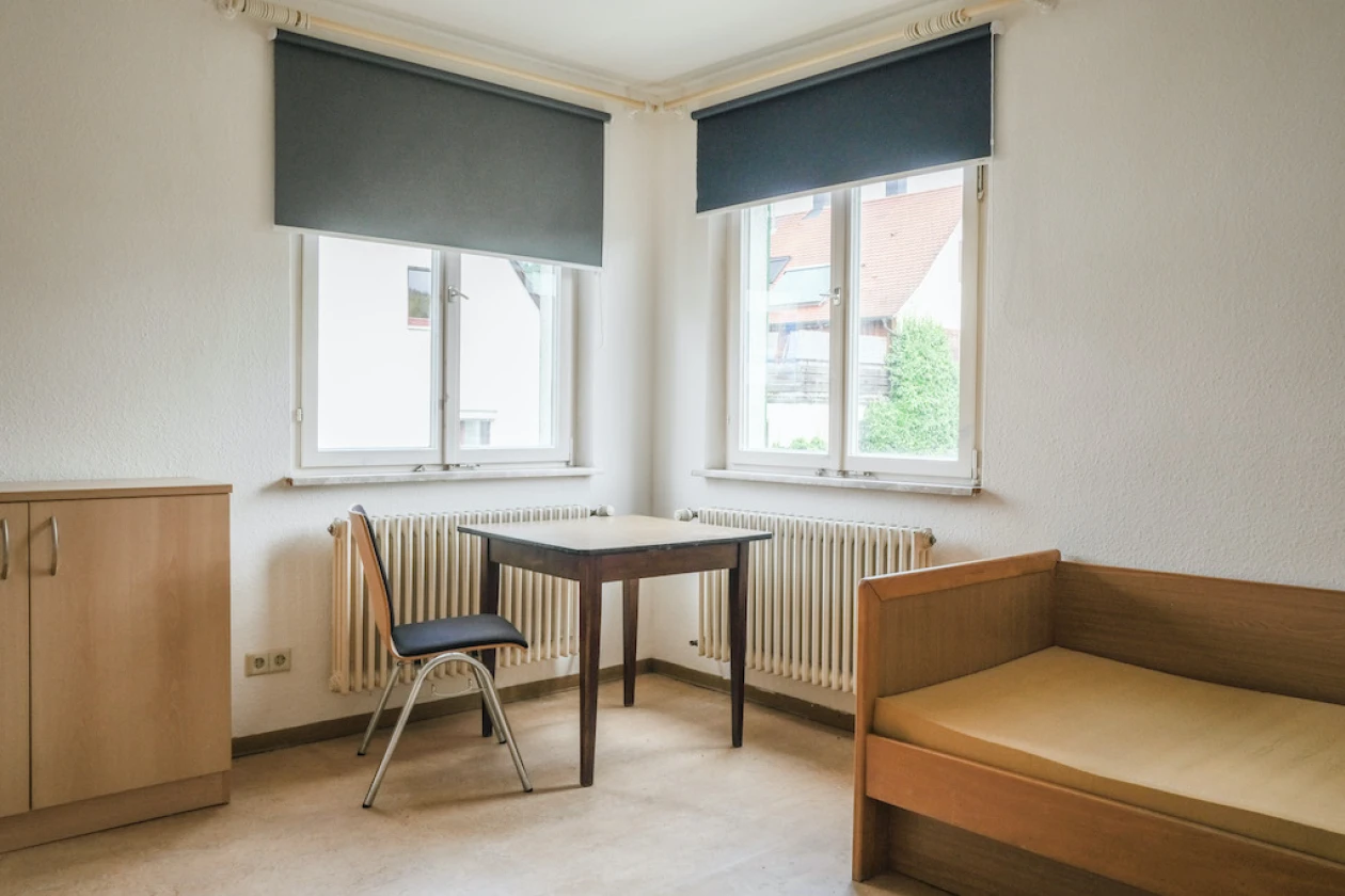 Ein Raum mit einem Einzelbett, einem kleinen Holztisch, einem Stuhl und zwei Fenstern mit Verdunkelungsrollos. Wei&szlig;e W&auml;nde, Holzm&ouml;bel und Heizk&ouml;rper unter den Fenstern pr&auml;gen den Raum.