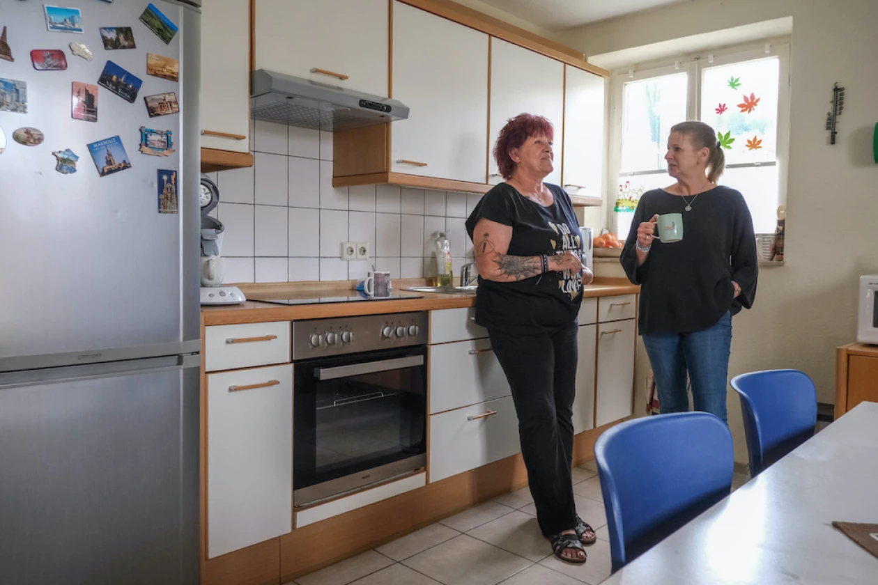 Zwei Frauen stehen in einer K&uuml;che und unterhalten sich, w&auml;hrend die rechte Frau eine Kaffeetasse in der Hand h&auml;lt. Im Hintergrund sind wei&szlig;e K&uuml;chenschr&auml;nke, ein Herd, ein K&uuml;hlschrank mit Fotos sowie ein herbstlich dekoriertes Fenster zu sehen.