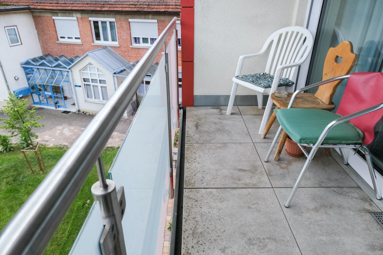 Balkon mit zwei Gartenst&uuml;hlen und einem Holzstuhl, Blick &uuml;ber das Gel&auml;nder auf ein Haus aus Klinkerstein mit wintergarten&auml;hnlichem Eingangsbereich und einen kleinen Rasenstreifen.