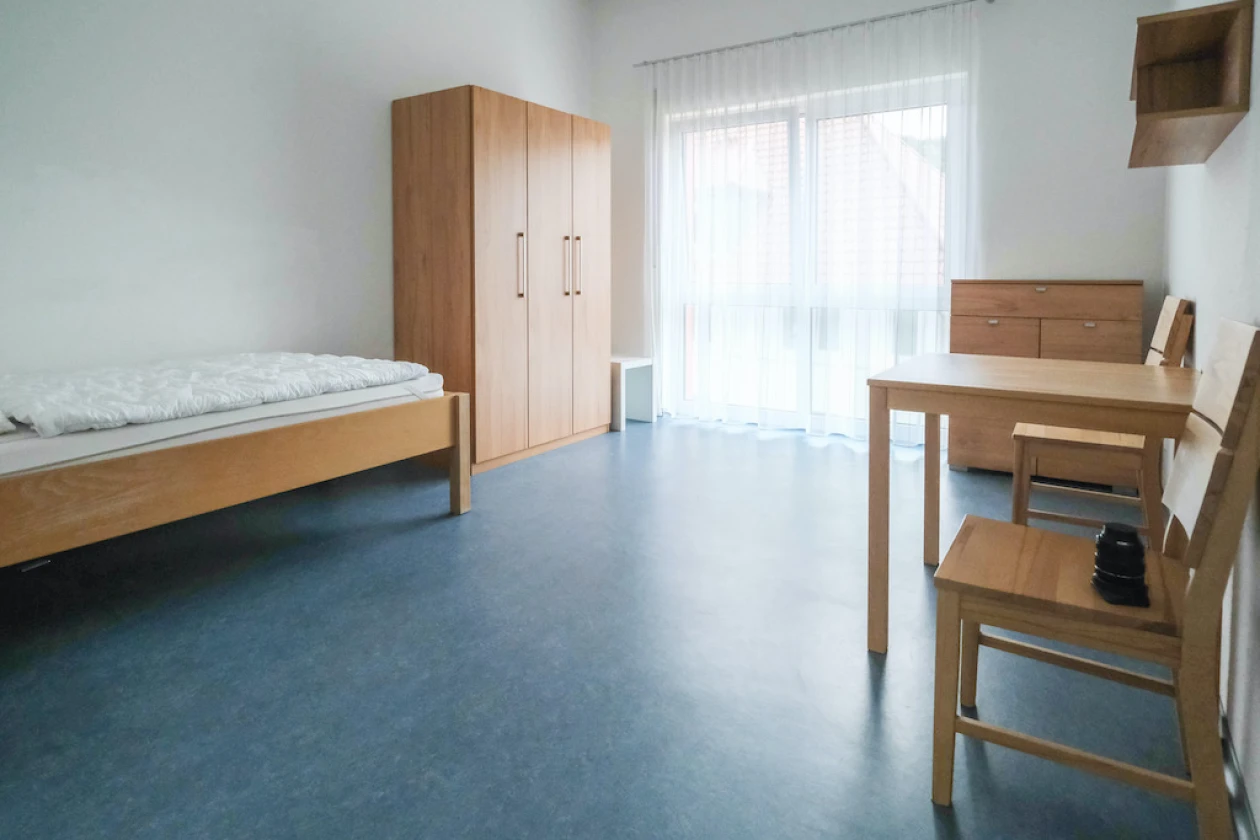 Ein Zimmer mit holzfarbenen M&ouml;beln: Ein Einzelbett, ein gro&szlig;er Schrank, ein Holztisch mit Stuhl und Fenster mit wei&szlig;em Vorhang. Der Raum hat wei&szlig;e W&auml;nde und einen blauen Bodenbelag.