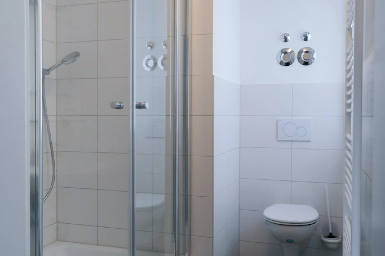 Ein Bad mit einer Glasduschkabine auf der linken Seite und einer Wandtoilette auf der rechten Seite. Wei&szlig;e W&auml;nde, graue Bodenfliesen und ein vertikaler Heizk&ouml;rper vervollst&auml;ndigen das Badezimmerdesign.