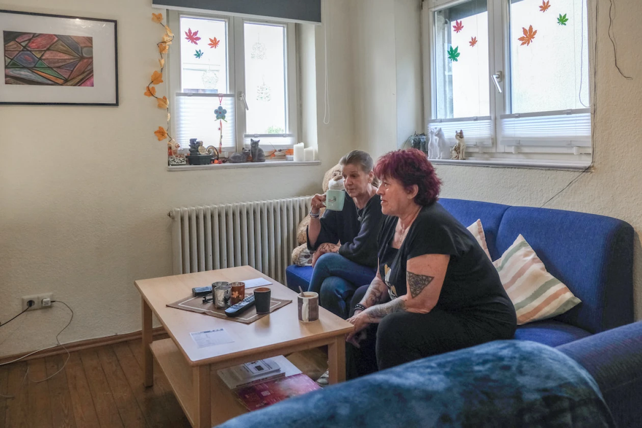 Zwei Frauen sitzen auf einem blauen Sofa an einem Holztisch. Im Hintergrund sind zwei Fenster zu sehen mit herbstlicher Dekoration und auf den Fensterb&auml;nken stehen kleine Figuren. Der Raum hat wei&szlig;e W&auml;nde, einen Heizk&ouml;rper und ein Gem&auml;lde an der Wand h&auml;ngen.