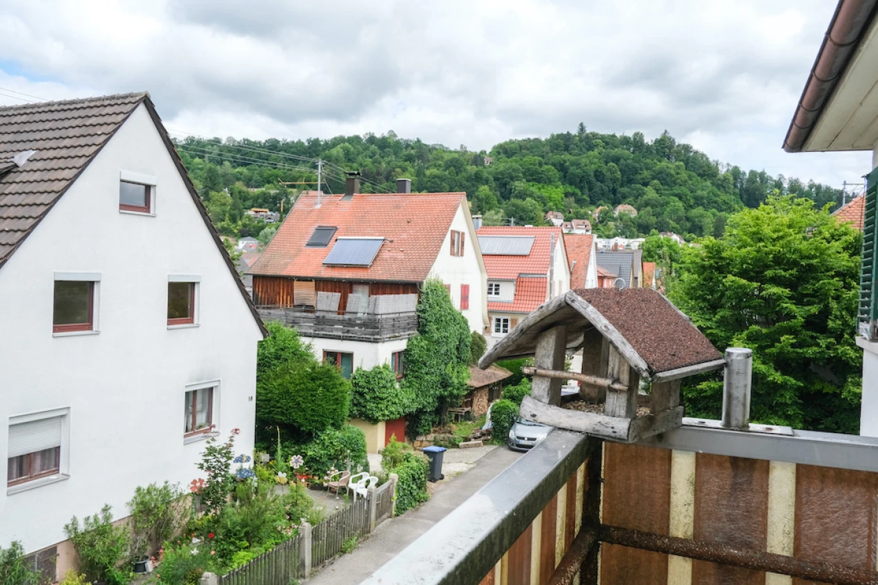 Blick von einem Balkon: Zu sehen sind mehrere H&auml;user, ein Auto und ein dichter Baumbestand, der an einen Wald erinnert. Auf dem Balkongel&auml;nder steht ein kleines Vogelhaus.